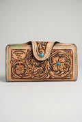 A hand-tooled brown leather wallet with floral engravings and a turquoise stone accent, resting on a textured tree trunk outdoors.