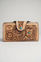 A hand-tooled brown leather wallet with floral engravings and a turquoise stone accent, resting on a textured tree trunk outdoors.