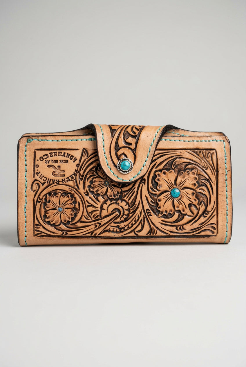 A hand-tooled brown leather wallet with floral engravings and a turquoise stone accent, resting on a textured tree trunk outdoors.